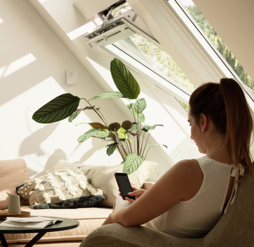 Frau beim Steuern ihrer VELUX Dachfenster in ihrem Wohnzimmer mit der VELUX ACTIVE App auf ihrem Smartphone.
