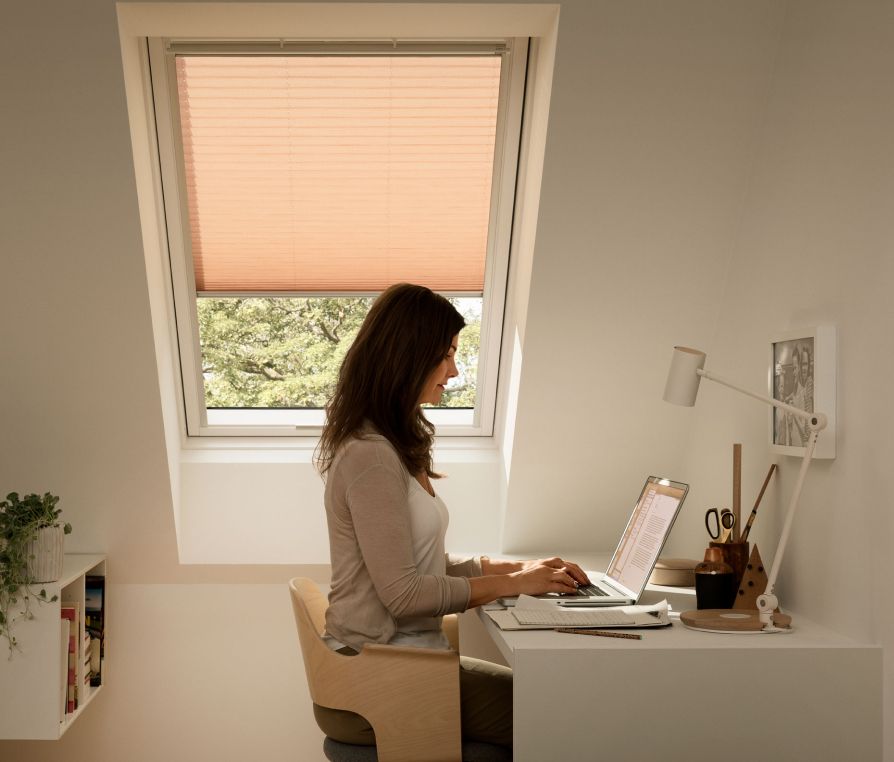 Frau in ihrem modernen Homeoffice, beim E-Mail-Schreiben auf ihrem Laptop mit einem VELUX Dachfenster im Hintergrund mit beiger Markisette.