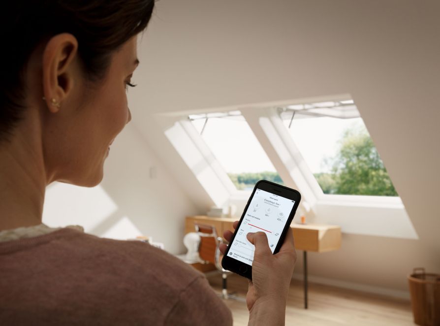 Une femme contrôle ses deux fenêtres de toit VELUX depuis son smartphone avec l'application VELUX ACTIVE with NETATMO.