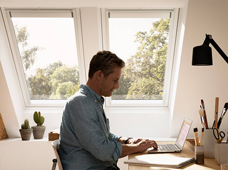 Ein Mann arbeitet an seinem Schreibtisch, während natürliches Licht durch zwei gestapelte VELUX Dachfenster strömt. 
