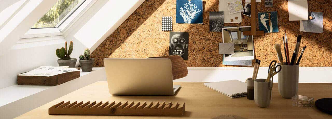 Schreibtisch mit Laptop, Lampe und Fotos an der Wand neben zwei VELUX Dachfenstern.