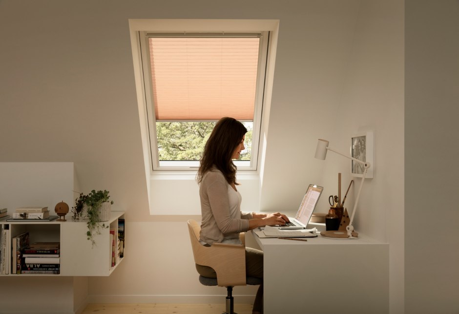 Eine an ihrem Tisch arbeitende Frau. Hinter ihr befindet sich ein VELUX Dachfenster mit einem bunten, heruntergezogenen Innenrollo.